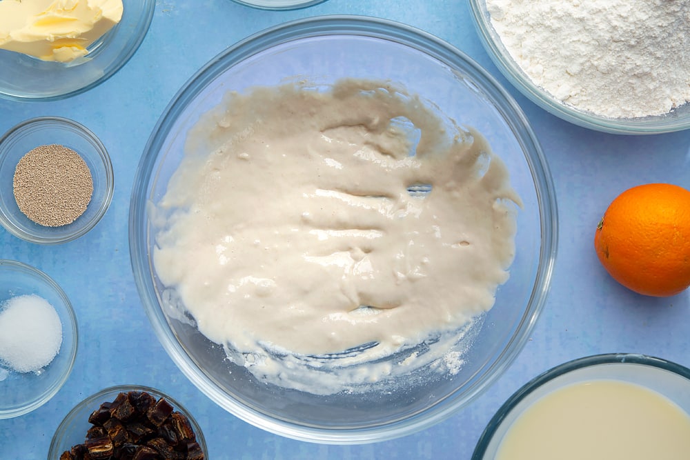 A bowl containing flour and water whisked together. The bowl is surrounded by the ingredients to make vegan hot cross buns.