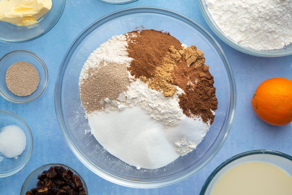 A bowl containing flour, yeast, sugar, salt, cinnamon, ginger and nutmeg. The bowl is surrounded by the ingredients to make vegan hot cross buns.