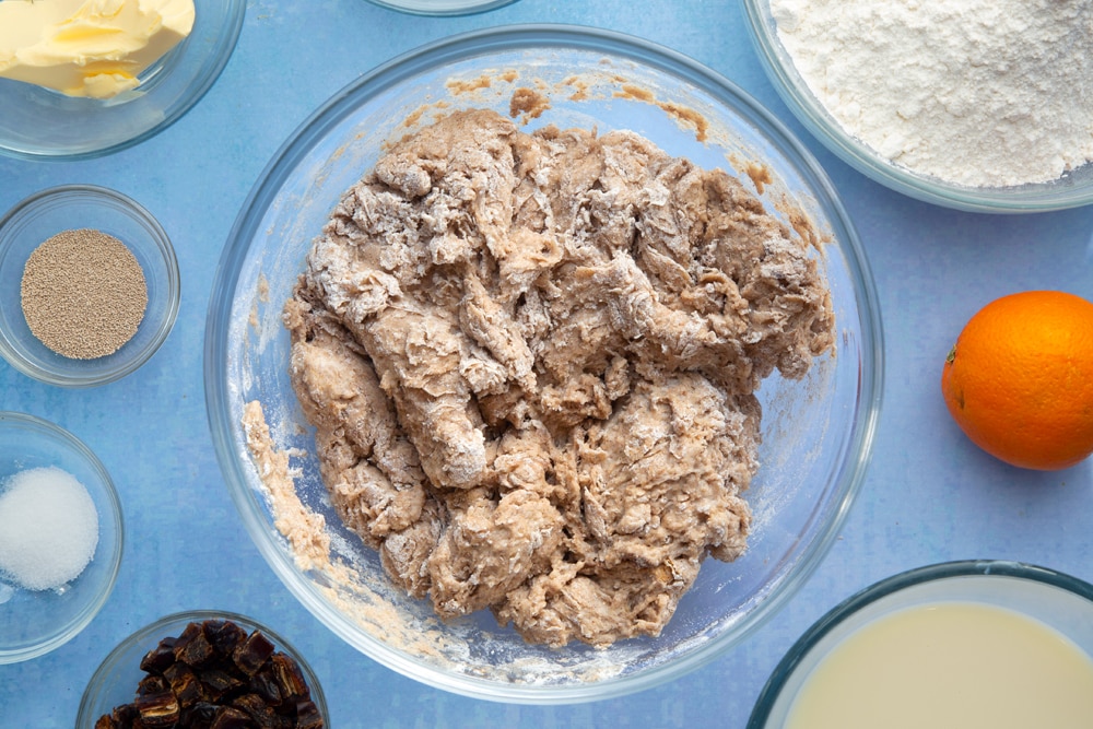 A bowl containing a roughly mixed spiced bread dough. The bowl is surrounded by the ingredients to make vegan hot cross buns.