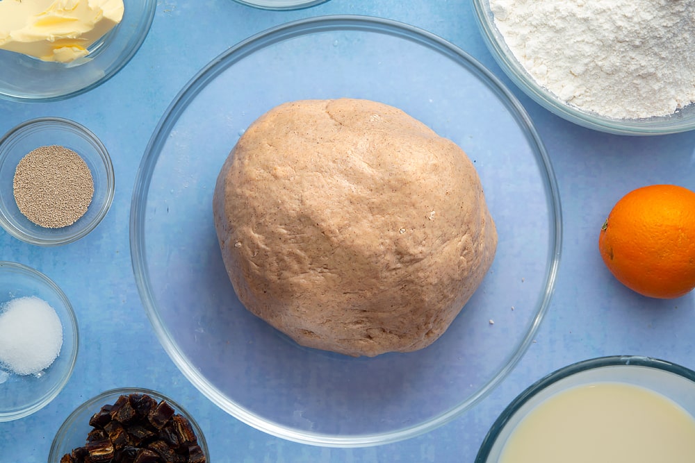 A bowl containing a smooth ball of vegan hot cross buns dough. The bowl is surrounded by vegan hot cross bun ingredients.