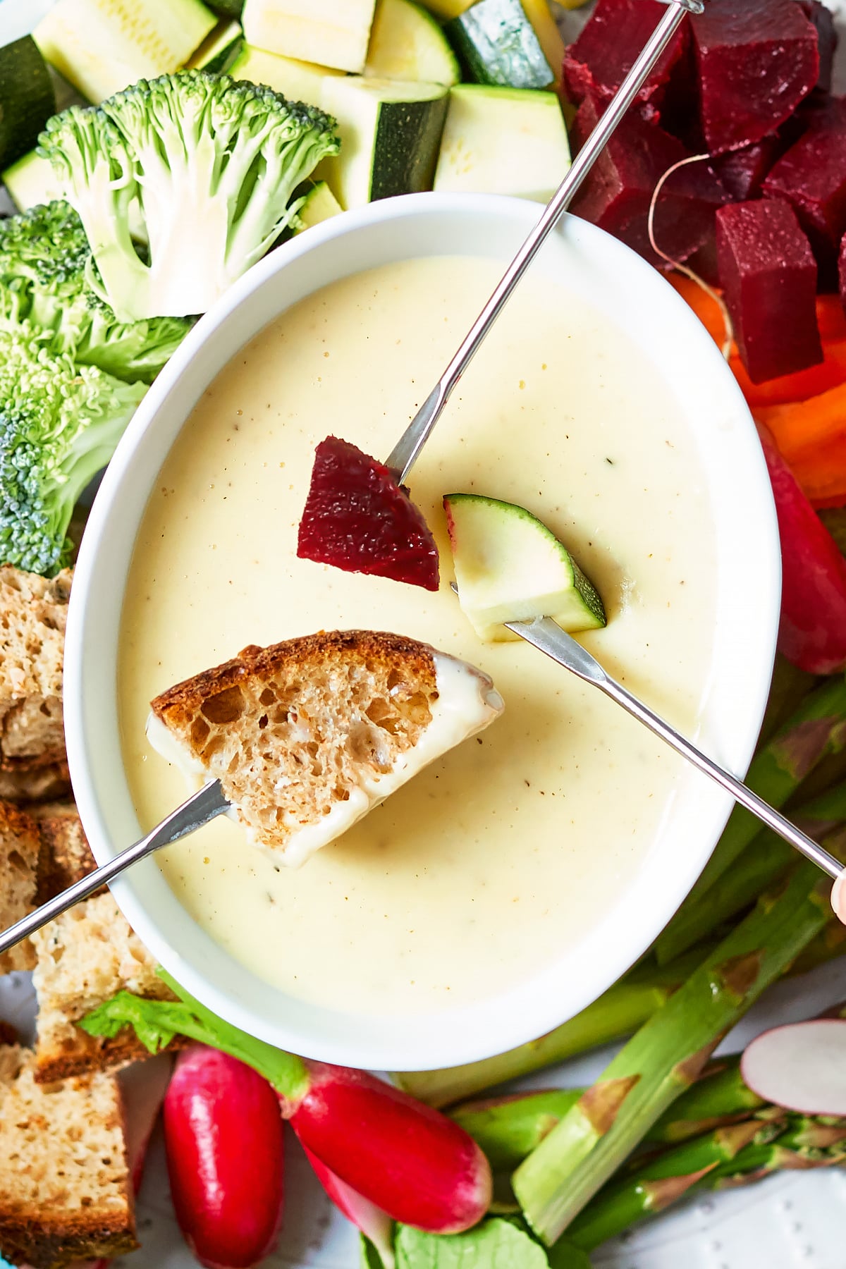 A close up of a fondue platter. Three fondue forks dip bread, beetroot, courgette into the cheese sauce.