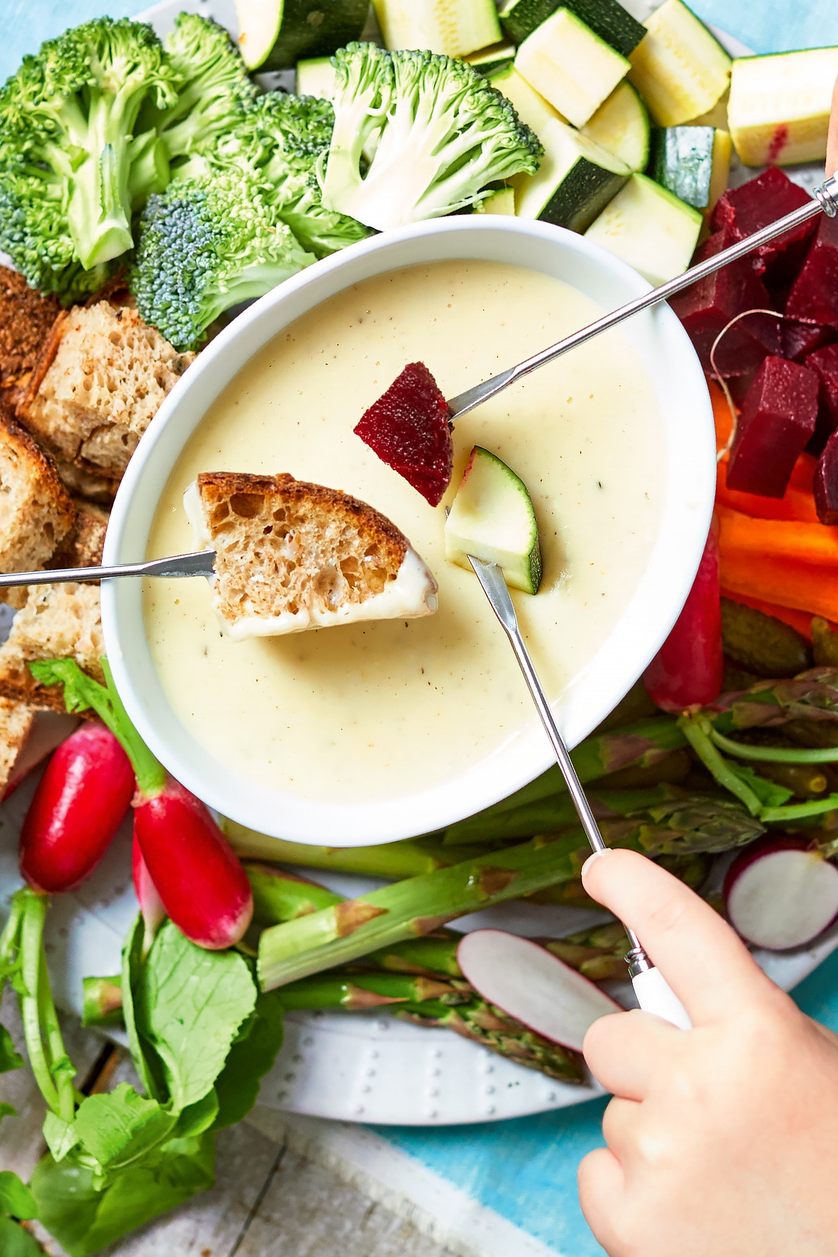 An overhead view of a vegetable fondue platter. A family of hands reach in with fondue forks to dip bread, beetroot, courgette into cheese sauce.