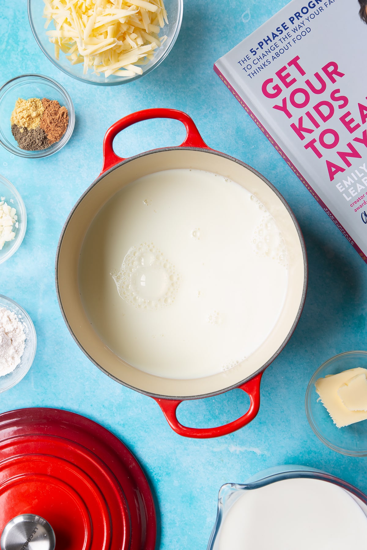 A pan containing milk. The pan is surrounded by ingredients to make cheese sauce for a vegetable fondue.