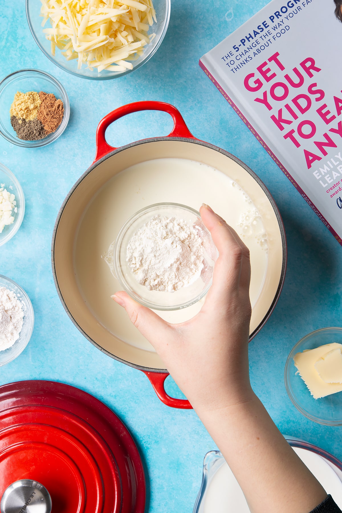 A pan containing milk. A hand holds a small bowl of flour. The pan is surrounded by ingredients to make cheese sauce for a vegetable fondue.