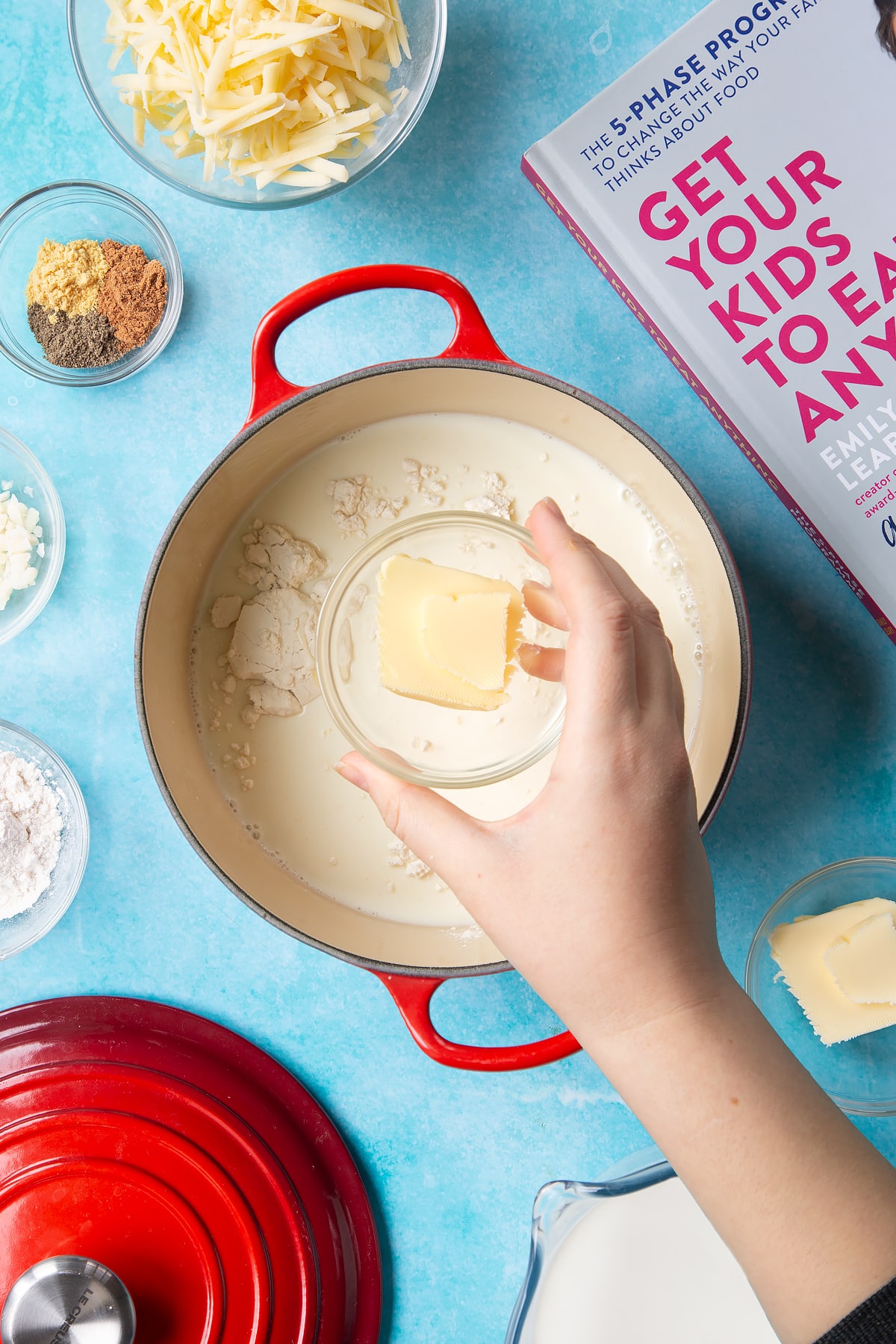 A pan containing milk, flour and garlic. A hand holds a small bowl of butter. The pan is surrounded by ingredients to make cheese sauce for a vegetable fondue.