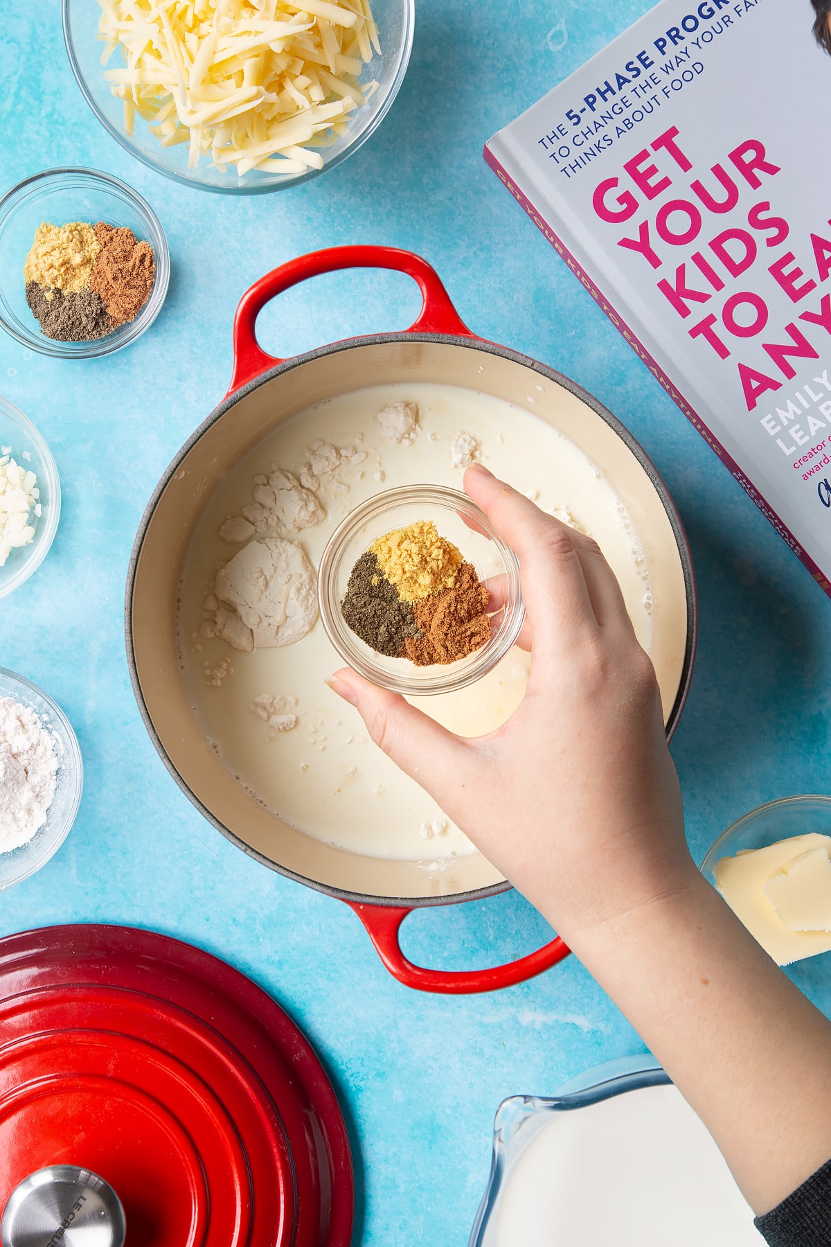 A pan containing milk, flour, garlic and butter. A hand holds a small bowl containing pepper, mustard and nutmeg. The pan is surrounded by ingredients to make cheese sauce for a vegetable fondue.