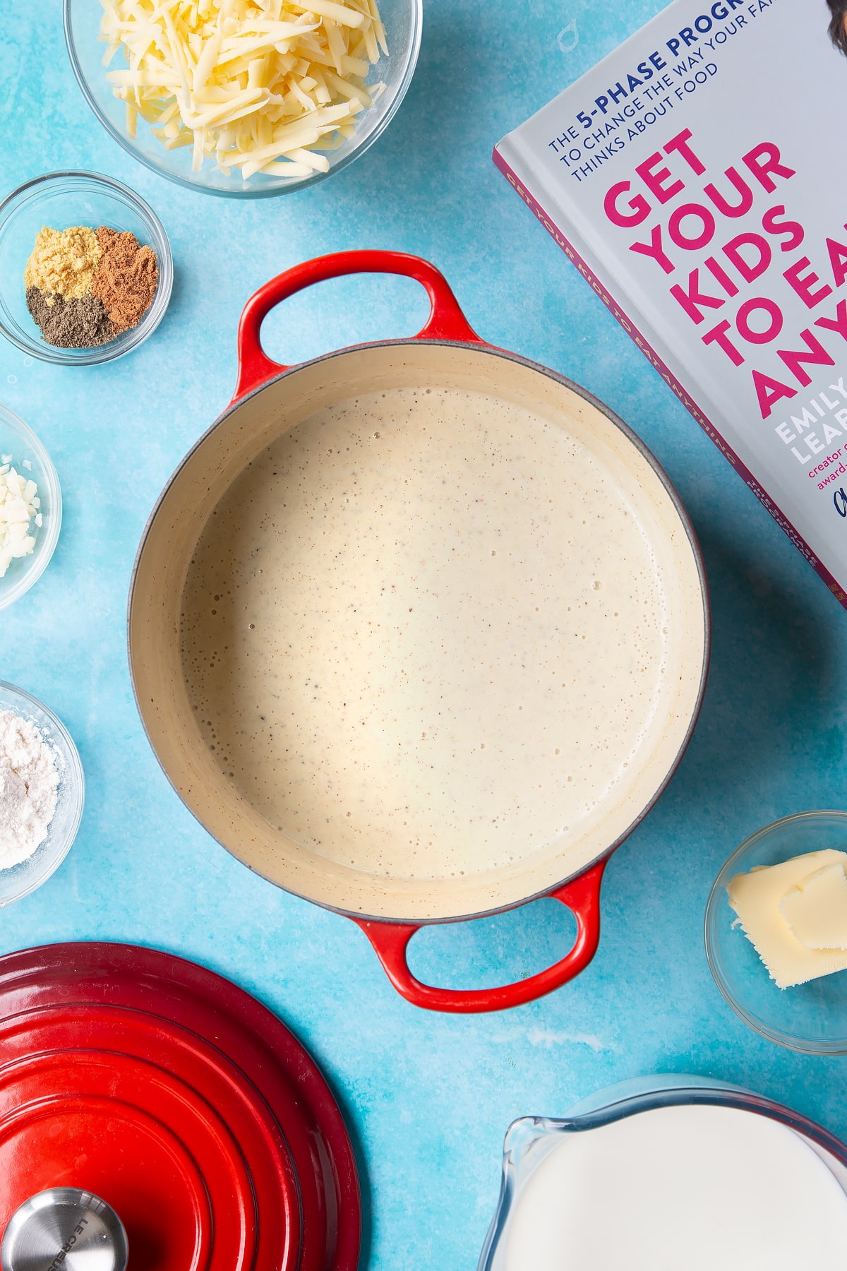 A pan containing a milk sauce, flavoured with garlic, pepper, mustard and nutmeg. The pan is surrounded by ingredients to make cheese sauce for a vegetable fondue.