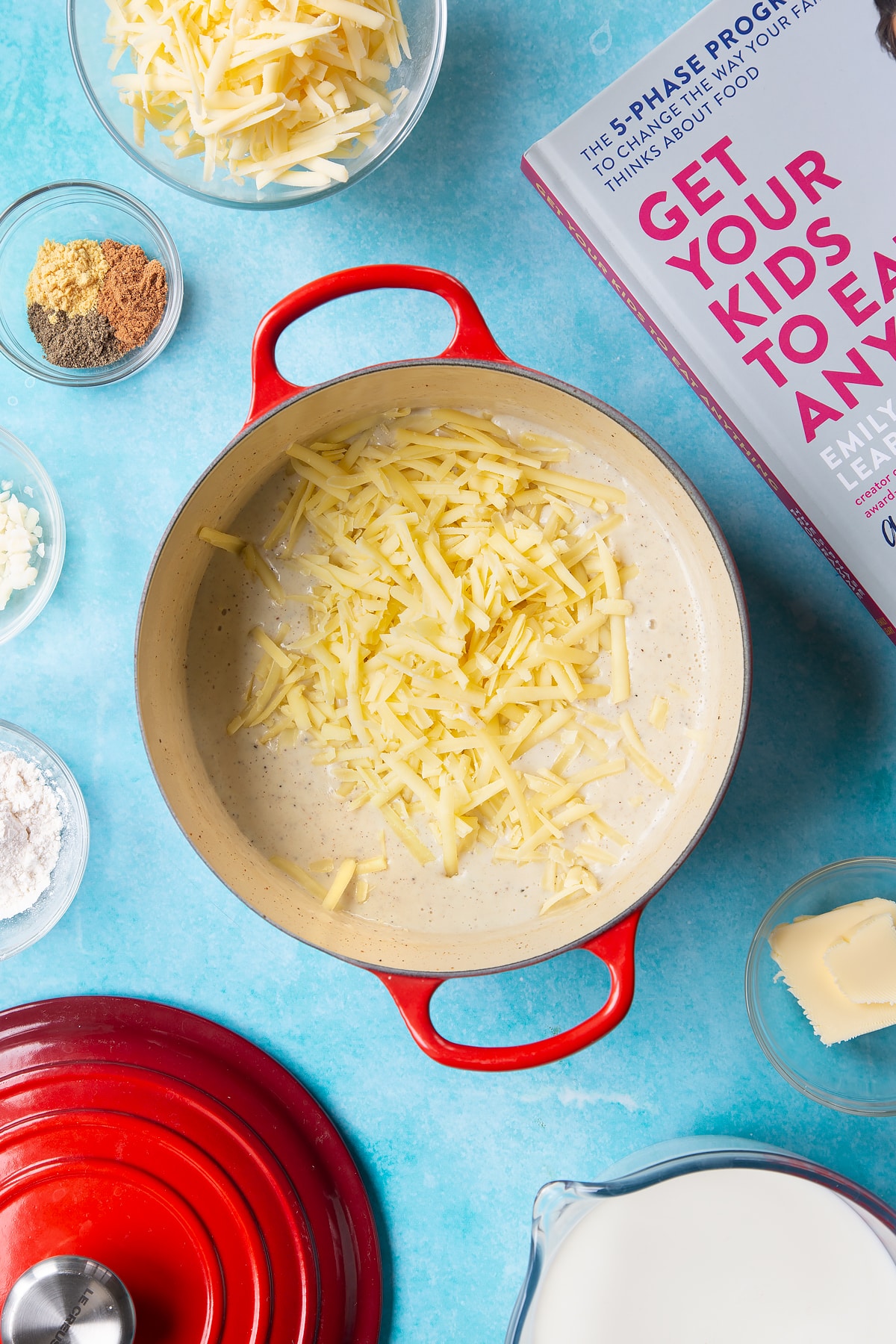 A pan containing grated cheese on top of a milk sauce, flavoured with garlic, pepper, mustard and nutmeg. The pan is surrounded by ingredients to make cheese sauce for a vegetable fondue.