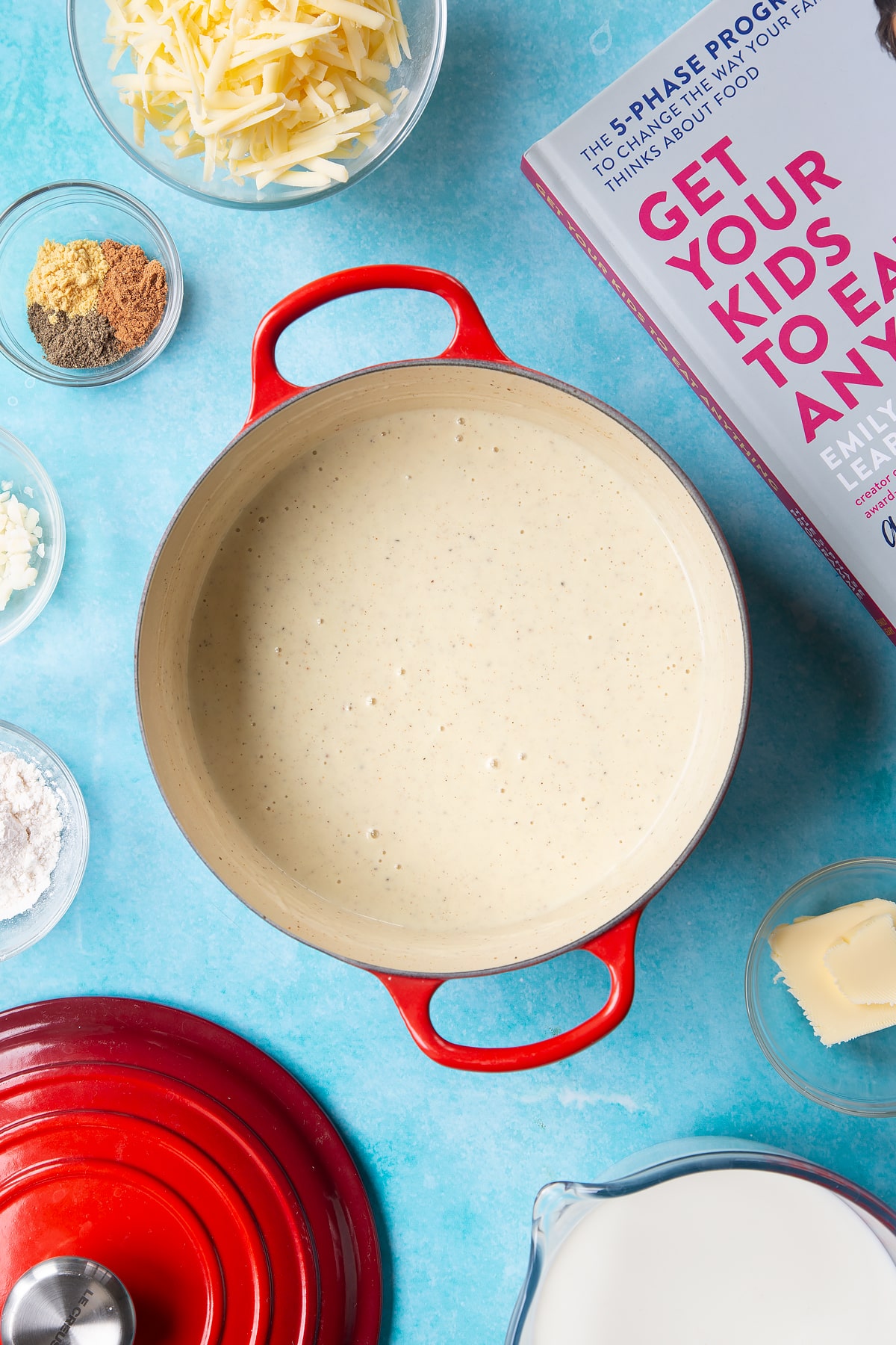 A pan containing a cheese sauce, flavoured with garlic, pepper, mustard and nutmeg. The pan is surrounded by ingredients to make cheese sauce for a vegetable fondue.